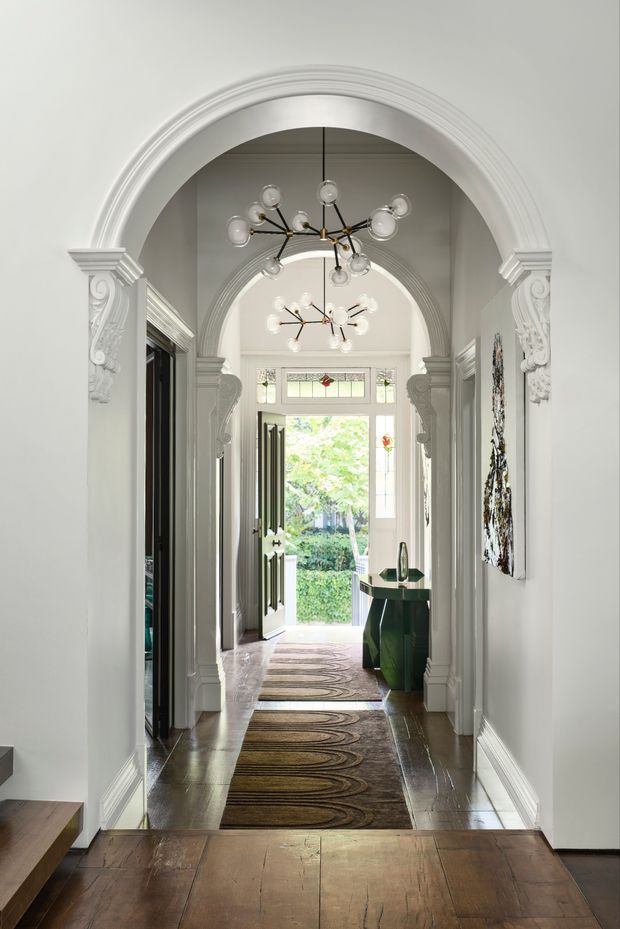 A refined residential hallway or entry corridor with a sequence of ornate white plaster arches, sculptural decorative corbels, dark timber flooring, and contemporary globe chandeliers. Patterned runners lead toward an open front door and a bright garden beyond. A dark green console table, artwork, and stained-glass transom detailing add character to the elegant heritage-style interior.
