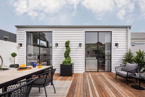 A contemporary outdoor timber deck and entertaining area adjoining a white horizontal-clad building. The deck features a large dining table with black woven chairs, a lounge chair, potted topiary and plants, and black-framed glazed doors opening into the interior. The scene is bright and refined with a warm timber and white palette.