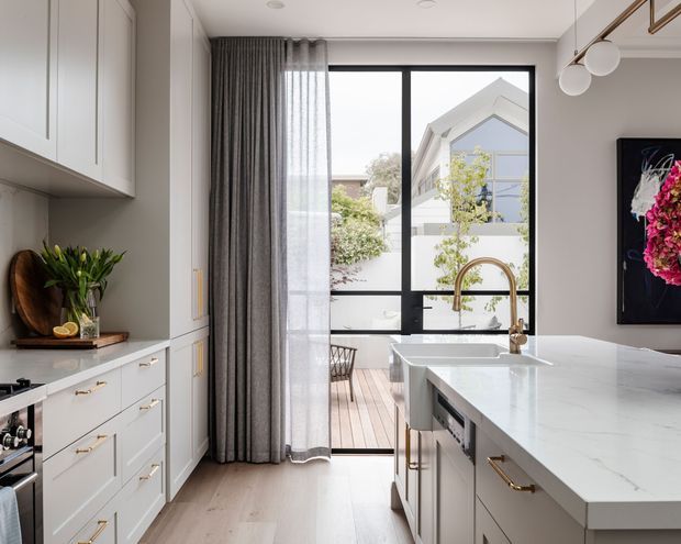 Bright, refined kitchen with white shaker-style cabinetry, brass hardware, a large white stone island, integrated appliances, and a brass gooseneck tap over a farmhouse-style sink. Full-height black-framed glazing opens to a timber deck, while layered grey curtains and a floral artwork add softness and colour.