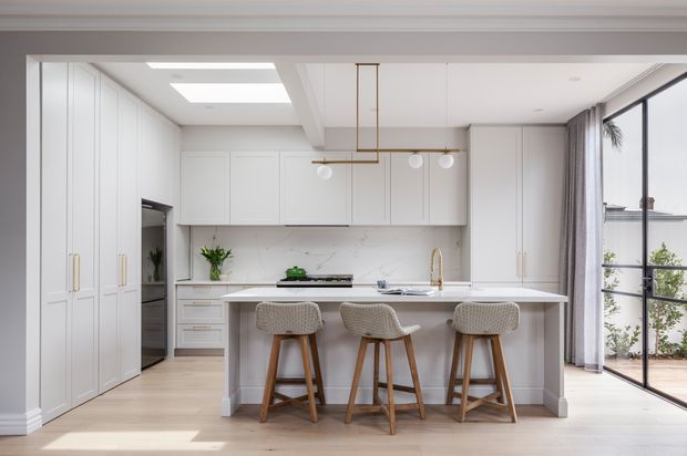 Bright, refined contemporary kitchen with extensive white built-in cabinetry, a large marble-look island with three woven bar stools, integrated appliances, skylights, and full-height glazing opening toward an outdoor area. Warm timber flooring and brass-toned hardware add subtle contrast.