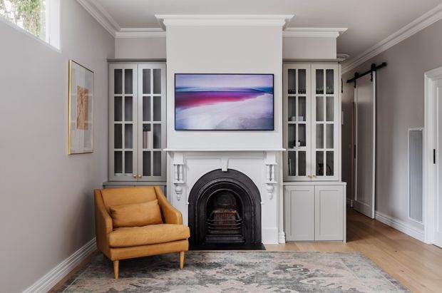 A refined living room with a white traditional fireplace and mantel, flanked by built-in glass-front cabinetry. The room has pale grey walls, white decorative trim and cabinetry, timber flooring, a patterned area rug, and a mustard leather armchair. A wall-mounted artwork or screen sits above the fireplace, while a black sliding barn-style door is visible to the right.