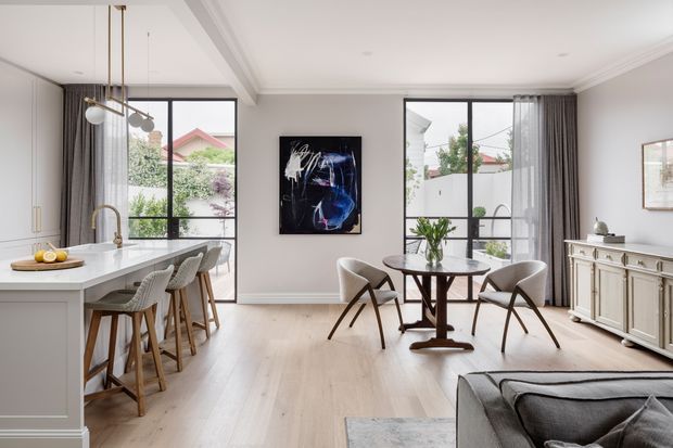Bright open-plan living and dining area with an integrated white kitchen island, pale timber flooring, black-framed floor-to-ceiling windows, a round dining table, contemporary chairs, a neutral sideboard, and abstract wall art. The space has refined light interiors with classic wall mouldings and a view to a leafy suburban setting.