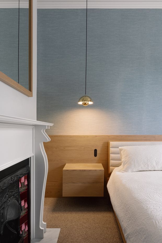 A refined bedroom interior featuring a white ornate fireplace surround, a timber half-height wall and floating bedside table, upholstered bedhead, soft blue-grey textured wall covering, carpeted floor, and a pendant light. The room combines traditional architectural detailing with contemporary timber and upholstered furnishings.