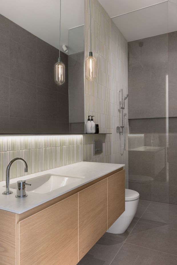 A contemporary bathroom featuring a floating light-wood vanity with an integrated white basin and chrome tapware. Large grey wall and floor tiles create a neutral backdrop, while a vertically ribbed pale tile feature wall continues behind the vanity and shower. A glass-enclosed shower, wall-mounted fixtures, under-shelf lighting, and suspended capsule pendant lights complete the refined design.