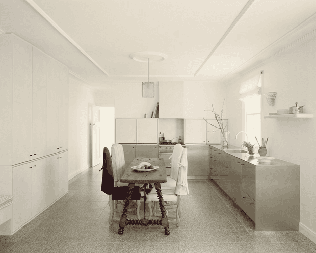 Bright, refined white kitchen-dining space with extensive built-in cabinetry, a large island, and a centrally positioned traditional dining table. The room features ornate ceiling cornices and decorative detailing, pale cabinetry, a grey terrazzo-like floor, pendant lighting, and understated neutral styling. A doorway and windows provide depth, while the island incorporates a sink and integrated storage.