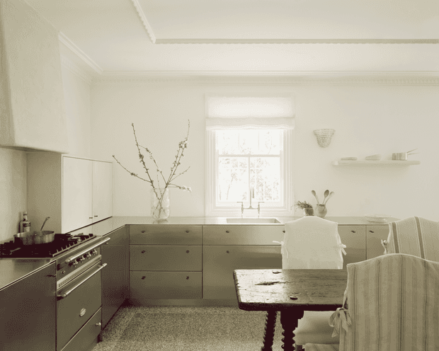 A bright, refined traditional kitchen and dining area with pale cabinetry, a large range cooker, stone-topped island or dining table, upholstered dining chairs, and a central window. Decorative ceiling moulding, a simple wall shelf, and understated accessories create a calm, classic interior.