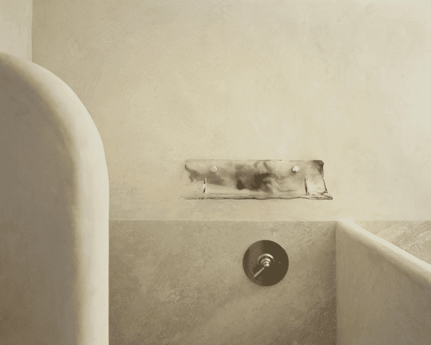 Close-up view of a sculptural, plastered bathroom or bathing area with softly rounded built-in forms, textured beige walls, a recessed metal plumbing plate, and a circular dark metal control or drain fitting. The muted monochromatic palette and curved surfaces create a minimalist, organic atmosphere.