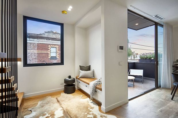 Bright contemporary apartment interior showing a compact living/sitting area with a built-in window seat, black round side tables, a large hide rug, and a black-framed window. A full-height glazed sliding door opens to a small balcony or terrace with outdoor seating. A vertical black slatted stair or screen element is visible at left, with light timber flooring and white walls throughout.