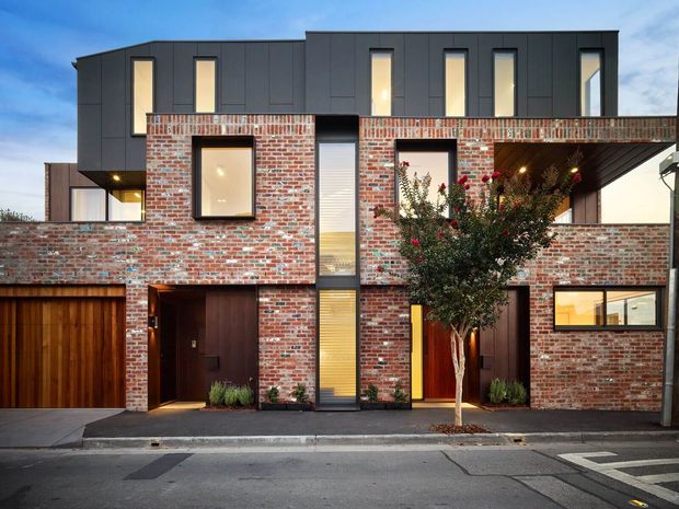 Street-facing view of a contemporary multi-storey urban townhouse development. The facade combines exposed red brick with dark charcoal upper cladding, tall narrow windows, recessed balconies, timber garage doors, and dark timber entry doors. A street tree and paved sidewalk sit in front of the building at dusk.