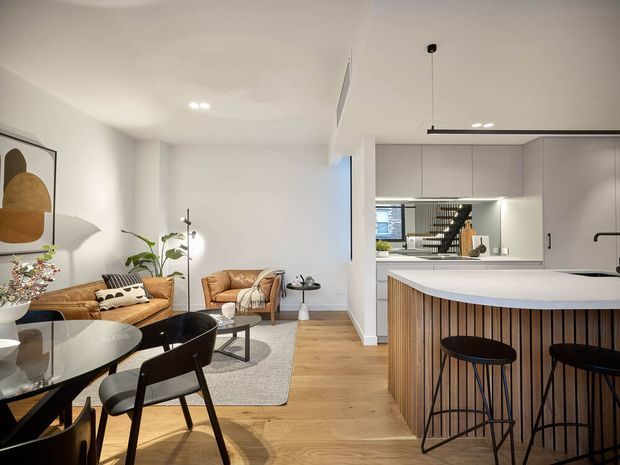 Open-plan contemporary kitchen, dining and living area with a curved white island and vertically slatted timber-clad base, light timber flooring, pale cabinetry, and integrated lighting. The adjacent living zone contains tan leather sofas, a round glass dining table with dark chairs, neutral textiles, and abstract wall art. A stairway and additional glazing are visible beyond the kitchen.
