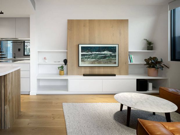 Bright contemporary living room with a wall-mounted television set against a vertical light timber feature panel, integrated white shelving and low cabinetry, light timber flooring, a pale textured rug, and an oval marble-look coffee table. A partial kitchen with timber island cabinetry is visible to the left, while leather seating and indoor plants add warmth.