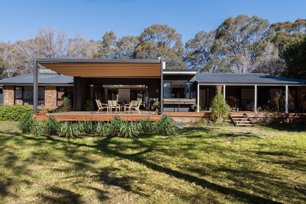 Single-storey residential house viewed from the garden, with a low-pitched dark roof, extensive glazing, brick walls, and a broad timber deck. A modern covered outdoor entertaining area with a timber-lined ceiling and dining furniture projects from the central living spaces, while established trees and drought-tolerant planting surround the lawn.