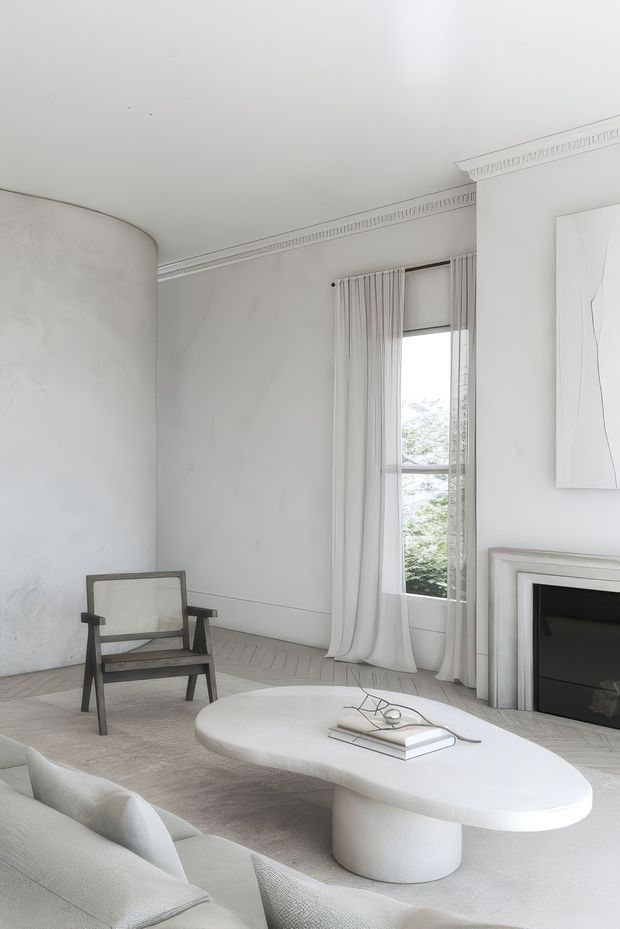 A bright, refined living room with a predominantly white and pale grey palette. The room features curved plastered walls, ornate linear cornice detailing, a tall curtained window, a classic fireplace surround with dark firebox, a low sculptural oval coffee table, a pale sofa, and a dark timber-framed armchair. The styling is quiet and minimalist with subtle classical architectural detailing.