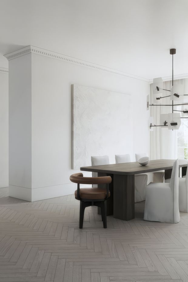 Bright, refined dining room with a dark timber dining table, upholstered white dining chairs, and a sculptural brown armchair. The room features pale herringbone timber flooring, white walls with decorative ceiling cornice detailing, a large abstract white wall panel, sheer curtains, and a contemporary suspended geometric pendant light.