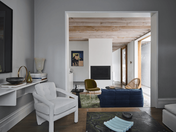Refined open-plan living room with a white upholstered armchair in the foreground, a deep blue sofa, olive accent chair, dark timber flooring, and a sculptural coffee table. A large framed opening connects the seating area to a secondary lounge with a wall-mounted fireplace, while full-height glazing and timber-framed doors bring in natural light. The ceiling features exposed pale timber boards and beams, contrasting with cool grey walls and white trim.