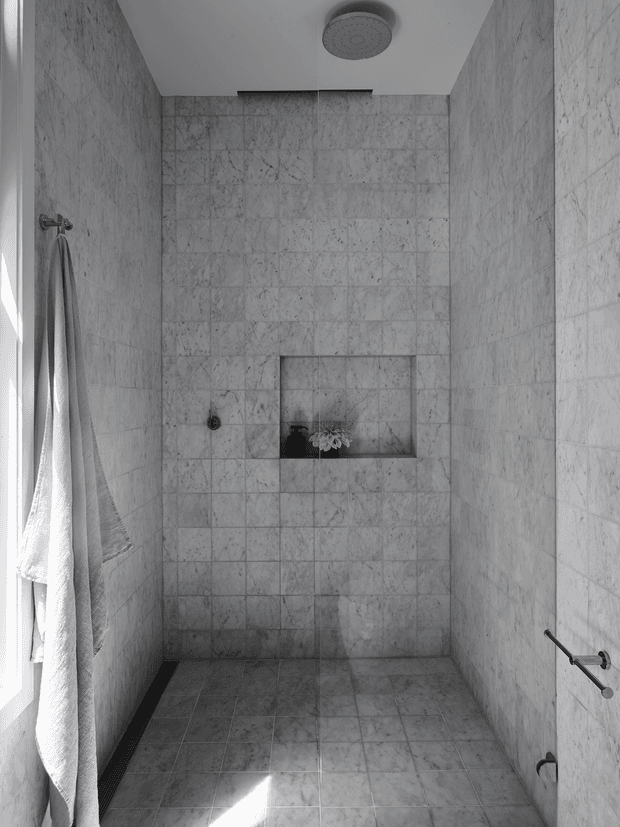 A compact walk-in shower with full-height grey-white veined stone or marble-look tiled walls and matching floor tiles. The shower has a ceiling-mounted rain shower, recessed wall niche, minimalist dark fixtures, and a towel hanging beside a bright window. The monochromatic space is clean, restrained, and naturally lit.