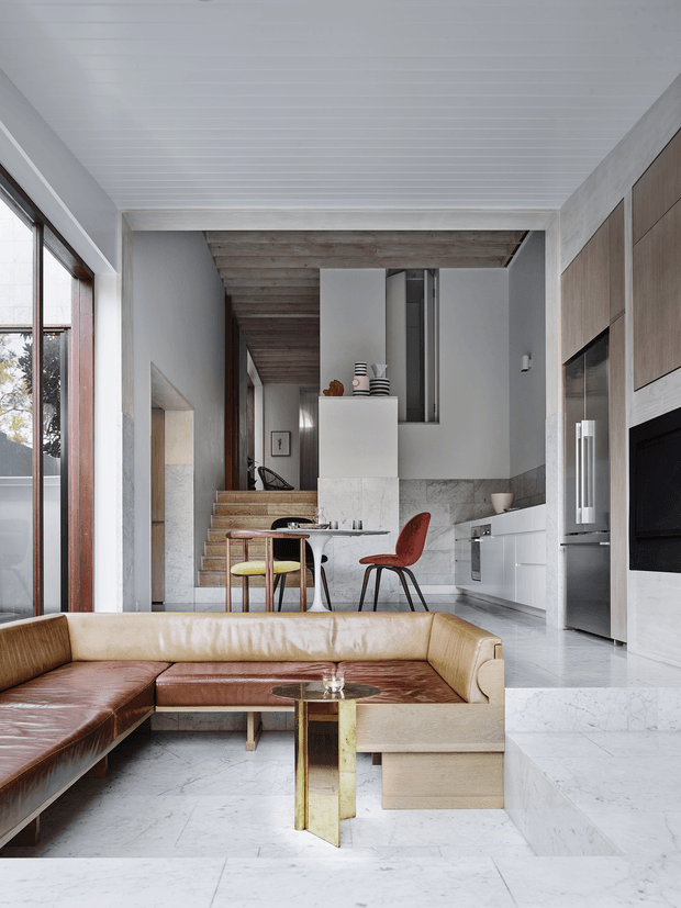 A refined open-plan living room and kitchen with a warm leather sectional in the foreground, a small round glass-and-brass coffee table, dining table and chairs, and white built-in kitchen cabinetry with integrated appliances. Pale marble or stone flooring and wall cladding contrast with dark timber ceiling beams, timber stair treads, and tall glazed openings. The space has a high, bright interior with a layered transition to a stairway and upper-level void.
