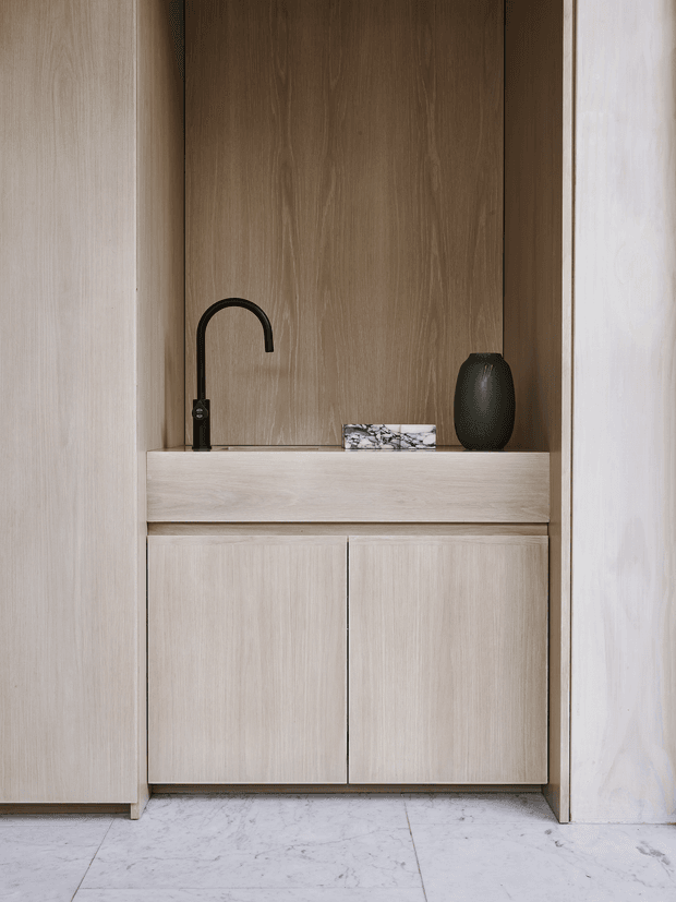A minimalist built-in timber vanity or washstand set within a recessed alcove. The light oak-toned cabinetry has two flat-front doors and a broad drawer or ledge, paired with a dark curved mixer tap, a small marble accessory, and a dark ceramic vessel. Pale marble-look floor tiles and light walls frame the warm wood joinery.