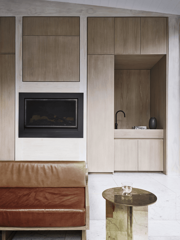 Refined contemporary living room featuring full-height light timber cabinetry and wall panelling, a black built-in fireplace, pale stone or marble flooring, and a warm brown leather sofa. A compact built-in bar or refreshment nook with a black tap and display vase occupies the right side, complemented by a round metallic coffee table. The composition has clean linear detailing, warm neutral tones, and an understated luxurious feel.