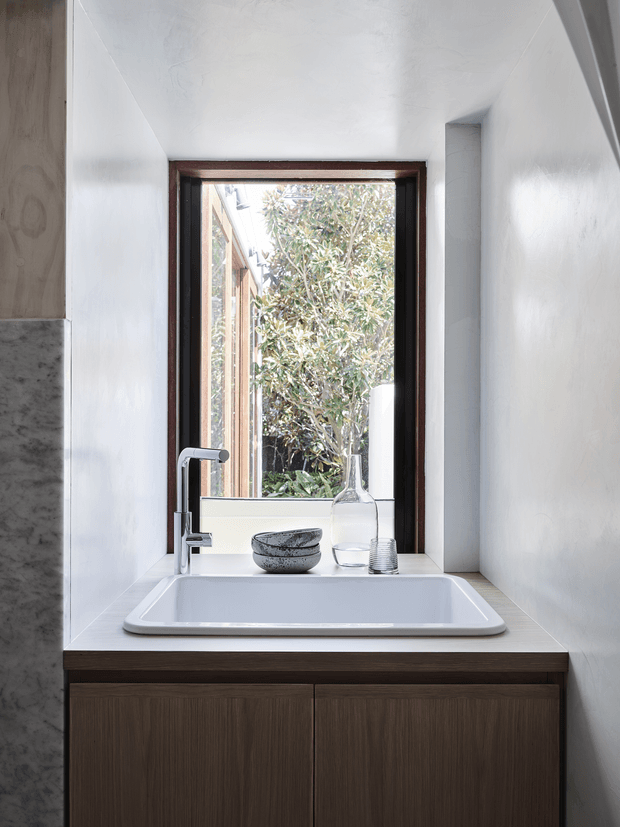Close-up view of a contemporary bathroom vanity beneath a timber-framed window. The white rectangular basin sits on a pale tiled benchtop above dark timber cabinetry, with a chrome tap and simple decorative vessels. White walls and abundant natural light create a clean, restrained composition.