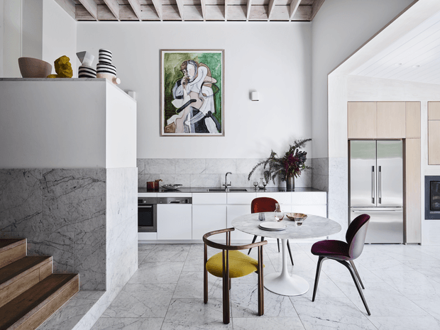 Bright contemporary kitchen and dining area with white cabinetry, integrated appliances, pale marble-look wall and floor surfaces, and a small round dining table with sculptural chairs. The room features an exposed timber-beam ceiling, an open stair edge, artwork, and a large opening into an adjacent space.