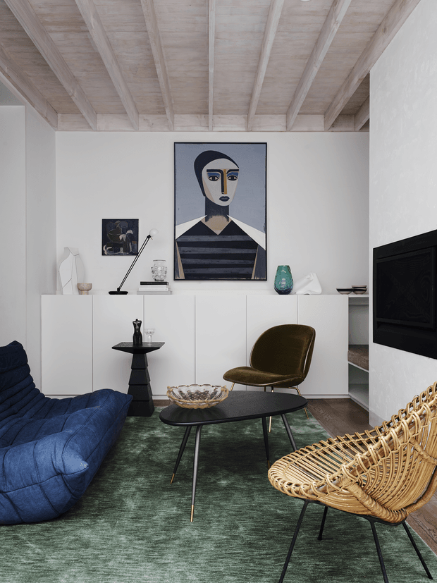 A stylish living room with a white built-in sideboard, dark oval coffee table, blue upholstered sofa, olive-green accent chair, and woven rattan lounge chair. The room features a pale timber ceiling with exposed beams, white textured walls, dark timber flooring, and a deep green rug. Mid-century-inspired artwork and decorative objects add visual interest, while a black wall-mounted fireplace or television feature anchors the right side.