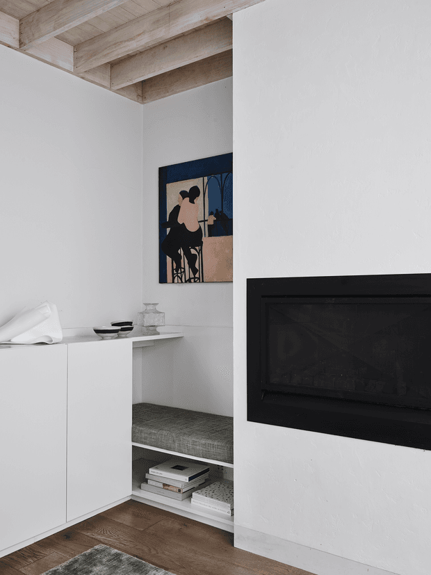 A minimalist living area with a built-in black fireplace set into a white plaster wall. White custom cabinetry and open shelving incorporate a grey upholstered window seat or bench, stacked books, ceramics, and a sculptural white object. A muted figurative artwork hangs in the niche, while exposed light timber ceiling beams and warm timber flooring add natural contrast.