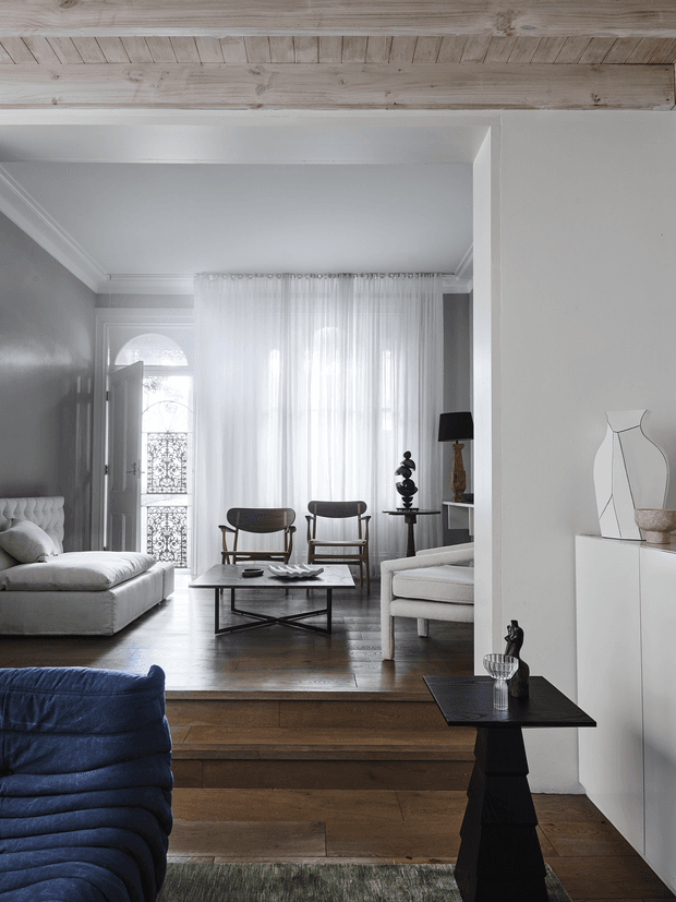 A refined, light-filled living room with a raised seating area and a timber-lined ceiling beam or soffit. The room features white upholstered sofas, mid-century-style wooden lounge chairs, a black rectangular coffee table, sheer full-height curtains, dark timber flooring, and a blue upholstered chair in the foreground. A sculptural white artwork and decorative objects sit on built-in white cabinetry, while an arched glazed entry door with ornate metalwork is visible at the rear.