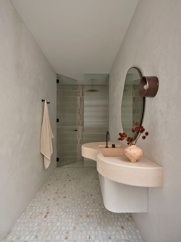 A narrow contemporary bathroom with softly textured off-white plaster walls and a curved floating vanity. The vanity includes an integrated pale stone or engineered-stone basin and dark metal tapware. A frameless glass shower enclosure with fine vertical ribbing sits at the rear, while the floor is finished in small irregular light mosaic tiles. An oval mirror, warm bronze wall light, and ceramic vase with dried stems add restrained decorative detail.