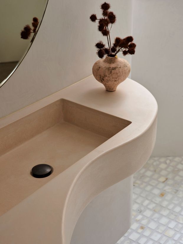 Close-up of a sculptural, rounded bathroom vanity with an integrated rectangular basin in a warm beige plaster or stone-like finish. A dark circular drain sits in the basin, while a textured ceramic vase with dried botanicals decorates the rear corner. A curved mirror is partially visible on the left, and the floor has small pale mosaic tiles. The scene has a soft, organic, minimalist character.