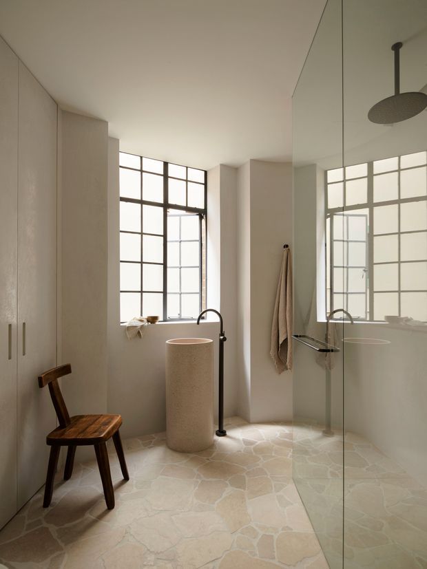 A serene, minimalist bathroom with a walk-in glass shower, freestanding round basin, wall-mounted showerhead, and freestanding bathtub visible through the glass. The room features pale plastered walls, large black-framed grid windows, a pale irregular stone floor, warm wood stool, and neutral linen towels. A full-height mirror reflects the shower and bathtub, enhancing the sense of space.