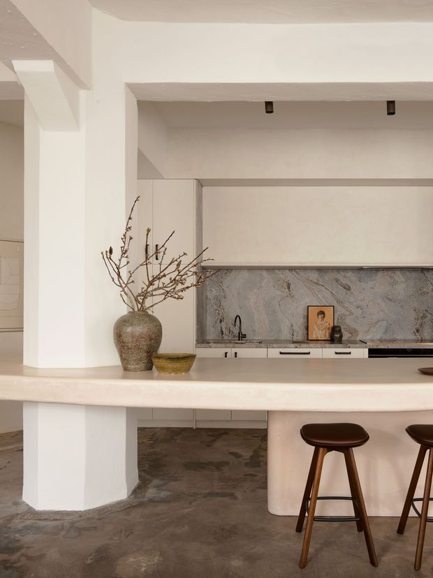 Warm, refined kitchen interior centered on a large curved light stone island with integrated seating. The space features pale built-in cabinetry, a dramatic grey veined stone splashback, dark mottled flooring, sculptural plasterwork, a minimalist sink and tap, and dark timber bar stools. Decorative branches and a ceramic vessel add an organic accent.