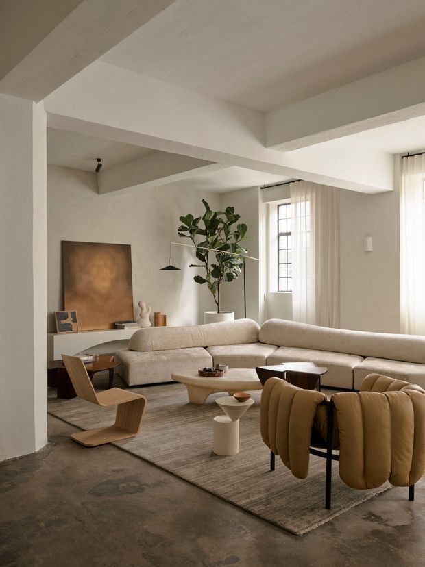 A softly lit contemporary living room with exposed structural ceiling beams, off-white plaster walls, tall steel-framed windows with sheer curtains, and a polished concrete floor. The room features a large cream sectional sofa, sculptural light-wood lounge chairs, rounded coffee tables, a sculptural pendant light, indoor greenery, and warm neutral furnishings arranged on a large textured rug.
