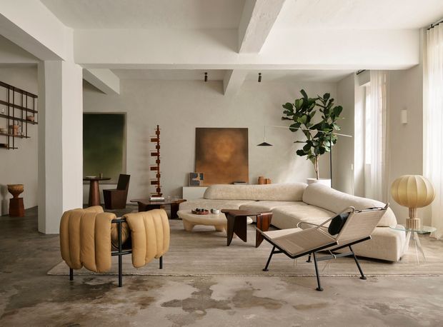 A spacious, refined living room with a cream modular sectional sofa, sculptural mid-century lounge chair, rounded upholstered armchairs, and low timber coffee tables. The room features exposed white structural beams, polished concrete flooring, neutral plaster walls, a large indoor fig tree, abstract artwork, open shelving, and generous sheer-curtained windows. The styling combines warm organic furnishings with restrained modernist architecture and soft natural light.