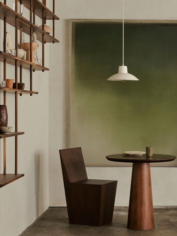 A refined dining nook with a dark timber pedestal table and sculptural timber chair, set against a large muted olive-green wall panel. Open vertical timber shelving displays ceramics and glassware along the left wall, while a simple white pendant light hangs above the table. The space has a warm, restrained, gallery-like atmosphere with an earthy palette and polished minimalist detailing.