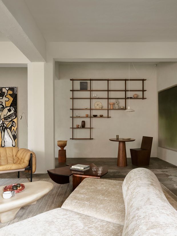 A warm, art-filled living room with cream plaster walls, exposed structural beams, dark stone flooring, and a large modular light-toned sofa in the foreground. A sculptural timber coffee table, rounded stone coffee table, leather lounge chair, small dining-style table, pendant light, and extensive open timber shelving with decorative objects create a refined contemporary interior. A large abstract artwork and wall-mounted dark screen are also visible.