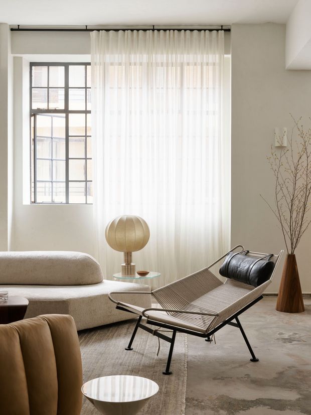 Bright, softly furnished living room with large industrial-style gridded windows, sheer full-height curtains on a black ceiling-mounted track, a cream curved sofa, sculptural sling chair, round floor lamp, glass side table, and a tall wooden vase with bare branches. The room has a neutral palette, polished concrete flooring, and a calm contemporary aesthetic.