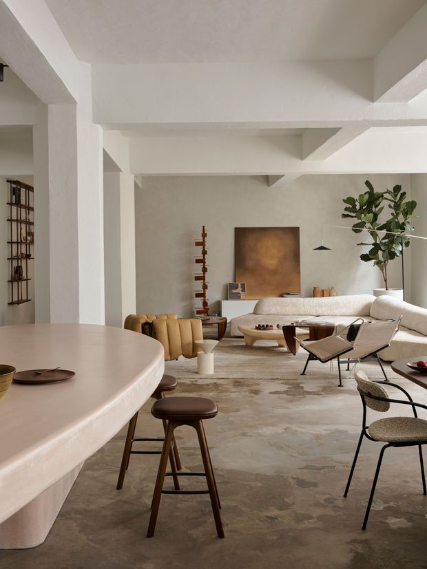 A spacious open-plan interior combining a dining area and living room. The room has white textured plaster walls and exposed structural beams, a large rounded pale stone or plaster dining island with wooden stools, a warm neutral sectional sofa, sculptural lounge chairs, a textured concrete-like floor, minimalist pendant lighting, and curated timber and ceramic furnishings. The palette is soft white, beige, taupe, and brown with a calm, refined contemporary character.