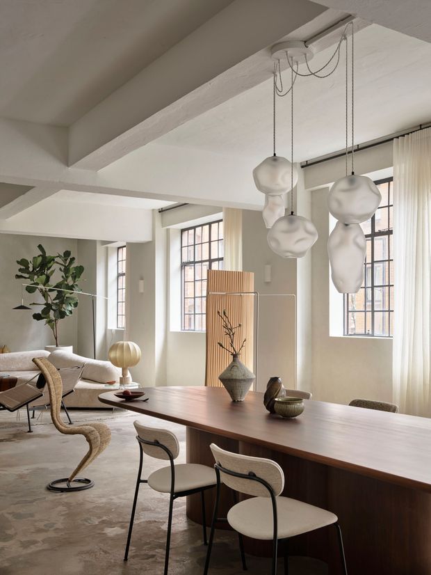 A refined open-plan dining and living interior with exposed white structural beams, tall gridded industrial-style windows, sheer curtains, and sculptural pendant lights. A long dark timber dining table with curved upholstered chairs occupies the foreground, while a neutral sofa, sculptural lounge chair, plants, and decorative objects create a warm contemporary setting.