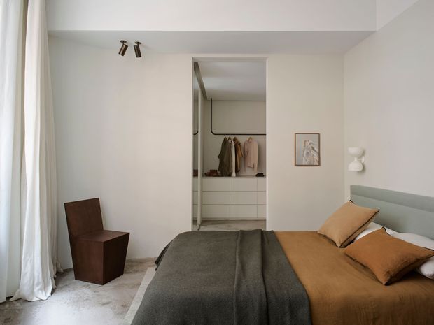 A calm, minimalist bedroom with a low upholstered bed, layered brown and charcoal bedding, and a simple brown accent chair beside full-height curtains. An open walk-in wardrobe is visible behind the bed, with white built-in drawers, hanging clothes, and a dark rail. The room has pale plaster walls, a light floor, a small framed artwork, a sculptural wall light, and discreet directional ceiling spotlights.