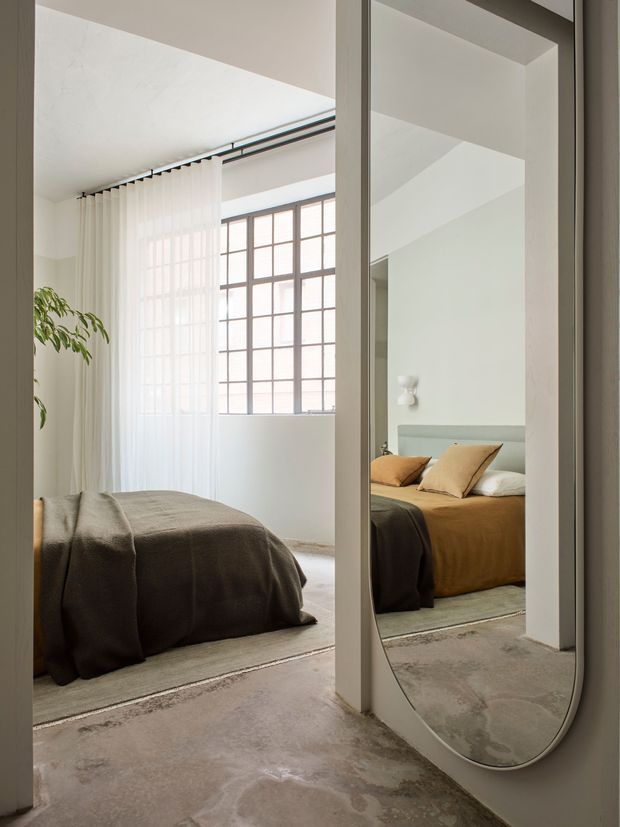 A bright, minimalist bedroom viewed through a full-height mirrored wardrobe or door. The room contains a low bed with muted taupe and ochre bedding, sheer white curtains, a large industrial-style gridded window, pale walls, and a raw concrete floor. The rounded mirror reflects the bed and emphasizes the room's light, spacious feel.