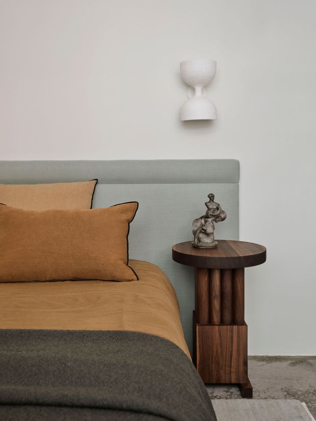 Close-up interior view of a softly styled bedroom corner. A muted grey upholstered bedhead sits against a warm off-white wall, layered with ochre-brown bedding and cushions and a dark grey throw. Beside the bed is a sculptural dark timber pedestal side table with a round top, topped by a small figurative sculpture. A minimalist white wall sconce is mounted above, creating a calm, refined, warm-toned composition.