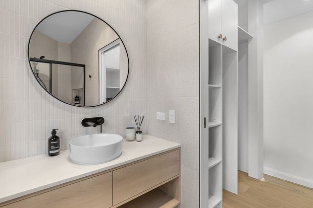 Bright contemporary bathroom featuring a floating light timber vanity with a white vessel basin, matte black wall-mounted tapware, a large round mirror, and pale vertically grooved wall tiles. Built-in open shelving and a white doorway lead toward an adjacent room with light timber flooring.