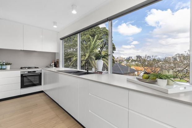 Bright contemporary white kitchen with extensive handleless cabinetry, a long peninsula or island with integrated sink, gas cooktop and built-in oven. Large corner windows provide abundant daylight and views of greenery, rooftops and a distant urban skyline. Light timber flooring and pale grey-beige benchtops create a clean, airy finish.