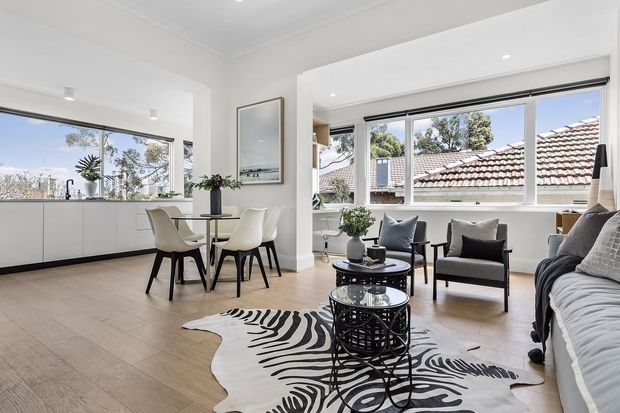 Bright, open-plan living and dining area with expansive corner windows, pale timber-look flooring, white built-in cabinetry, a compact dining table, lounge seating, nested round coffee tables, and a bold black-and-white patterned hide rug. The room has abundant natural light and views toward neighbouring tiled roofs and trees.