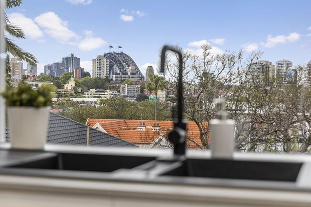 View from a kitchen sink through a large window toward a leafy urban neighbourhood and city skyline. The foreground shows a dark double-bowl sink, tall black gooseneck tap, pale benchtop, and potted plants, while terracotta rooftops, mature trees, high-rise buildings, and a distinctive arched bridge structure are visible outside.