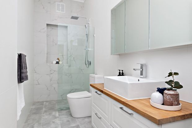 Bright contemporary bathroom featuring a frameless glass walk-in shower lined with white marble-look wall tiles, grey stone-look floor tiles, a wall-hung toilet, and a white vessel basin on a timber benchtop with white built-in vanity cabinetry. Mirrored wall cabinetry and minimal black accessories complete the clean scheme.