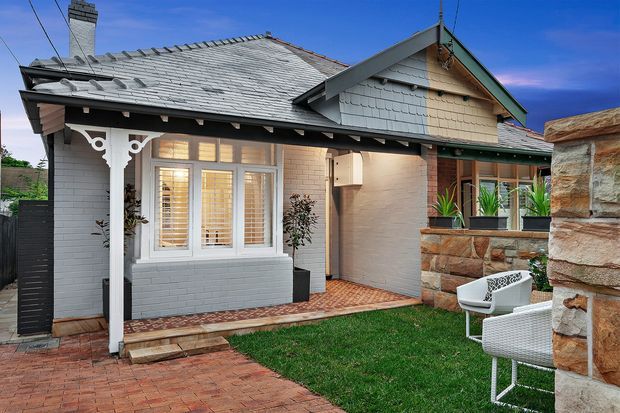 Exterior view of a single-storey traditional bungalow with a grey tiled gabled roof, painted brick walls, decorative white timber porch posts, and a covered entry patio. The landscaped courtyard includes brick paving, lawn, raised stone garden walls, outdoor seating, and potted plants.