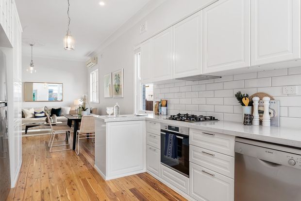 Bright white kitchen with shaker-style cabinetry, a white tiled backsplash, stone-look benchtops, stainless-steel appliances, and warm timber flooring. The open-plan layout connects directly to a dining and living area, with pendant lighting and understated coastal-traditional detailing.