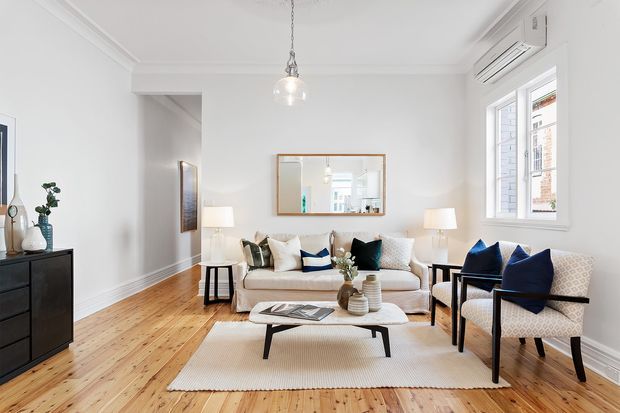 Bright, refined living room with white walls and decorative ceiling mouldings, warm timber floorboards, a cream sofa, patterned armchairs, a black-framed coffee table, lamps, and a neutral rug. Large windows and a wall-mounted air conditioner provide abundant natural light, while a framed mirror and curated accessories add polish.